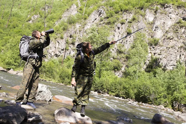 The forester and the inspector with a backpack, sleeping mat behind his back and a rifle in the trek across the mountain river, flowing among the stones in the forest