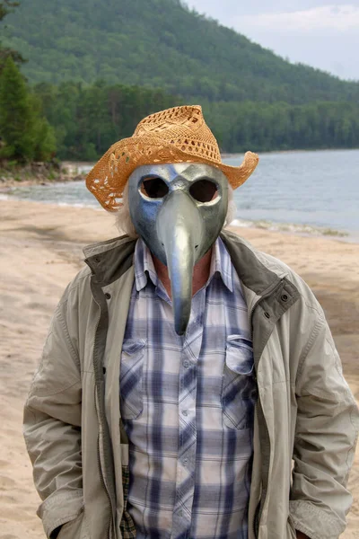 Senior man in a hat and a plague mask walks in cloudy weather along the shore of Lake Baikal