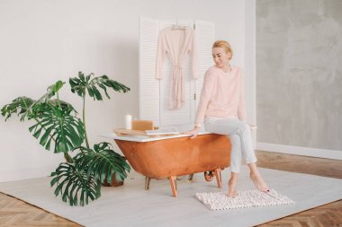 Genç bir sarışın pijamalarıyla banyonun kenarında çıplak ayakla oturmuş düşünceli bir şekilde gülümsüyor.