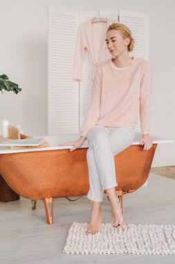 Genç bir sarışın pijamalarıyla banyonun kenarında çıplak ayakla oturmuş düşünceli bir şekilde gülümsüyor.