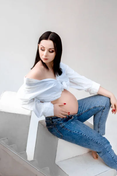 Vacker ung gravid kvinna brunett i blå jeans kramar magen på vit bakgrund — Stockfoto