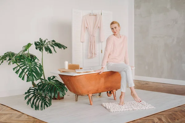 Genç bir sarışın pijamalarıyla banyonun kenarında çıplak ayakla oturmuş düşünceli bir şekilde gülümsüyor.