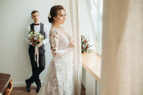 Düğün. Damat elinde bir buket tutuyor ve geline bakıyor. Ön plandaki gelin pencereden dışarı bakıyor.