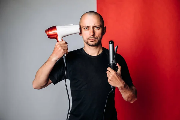 a bald brutal man, holding a hair dryer and Curling tongs in his hand, dries his hair and baldness . A man in a black t-shirt on a red and gray background. Hair care concept. male hairdresser