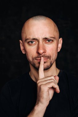 Genç ve kel bir adamın portresi, kameraya bakıyor ve ağzına bir parmak bastırıyor, o da sessizlik istiyor. - Korku konuşma korkusudur. Siyah arkaplana karşı siyah bir tişörtle