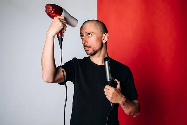a bald brutal man, holding a hair dryer and Curling tongs in his hand, dries his hair and baldness . A man in a black t-shirt on a red and gray background. Space for text. Hair care concept