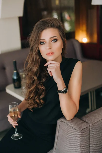 Romantic sad woman with long hair holds glass of champagne. Gorgeous long healthy hair. Brown-hairedmodel with waving hair in black. Professional makeup, sexy sensual lips