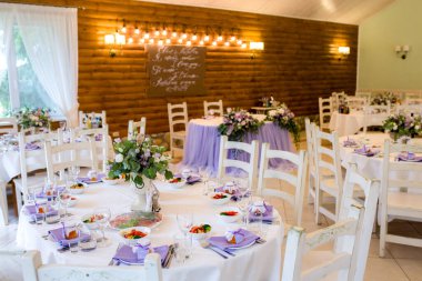 Beyaz güller, yeşillik ve mor çiçeklerden oluşan güzel tasarlanmış bir düğün masası ve arka planda ampullerden oluşan ahşap bir çelenk. Mor renk şeması.