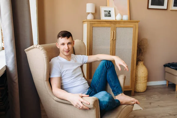 A young guy is sitting in a beige armchair barefoot and looking at the camera. Casual style, blue jeans and a white T-shirt. Stay indoors. Dream enjoy life. beige chair, a light and stylish interior.