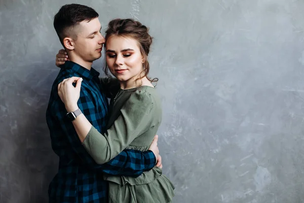 Handsome guy hugs his girlfriend. Happy Cheerful Family. Valentine\'s Day. Posing against a gray wall. Hugging. a girl in a khaki dress, he has a green plaid shirt. place for text
