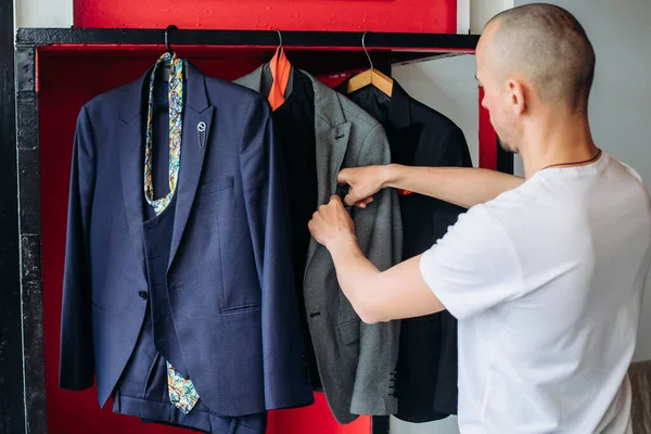 In the face of difficult choices. A bald young man makes a choice of a suit for a job. The guy chooses which official costume to wear. Choice of clothes. a man holds jackets in his hands at home