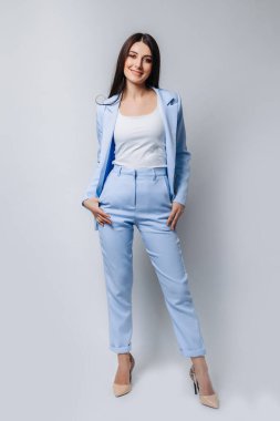 Düz kesimli esmer saçlı, resmi mavi takım elbiseli ve bej topuklu ayakkabılı bir iş kadını. Gülümsüyor. Gri arkaplanda tam vücut uzunluğu izole edildi