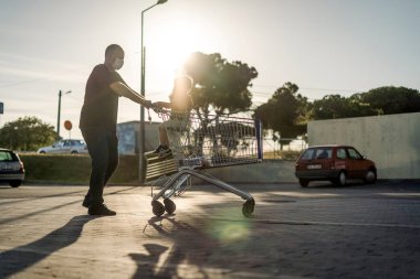 Baba, oğluyla birlikte bir otoparkta alışveriş arabasını itiyor. İkisi de koruyucu maske takıyor..