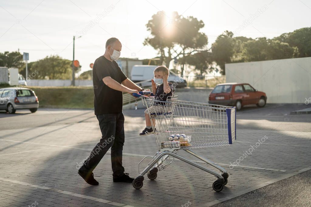 Father pushing shopping cart with his little son on a parking lot. Both wearing protective masks.