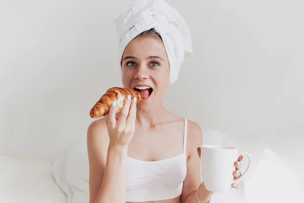 Wanita lucu bergairah dengan handuk di kepala menggigit croissant pagi dan minum kopi di tempat tidur putih — Stok Foto