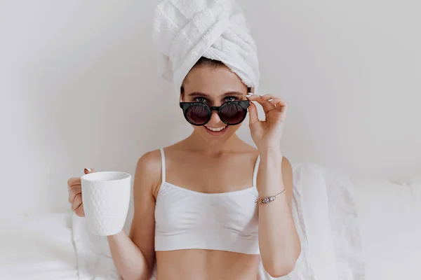 ผู้หญิงแฟชั่นที่มีผ้าเช็ดตัวรอบหัวของเธอสวมเสื้อสีขาวถือถ้วยและดูผ่านแว่นตากันแดด — ภาพถ่ายสต็อก