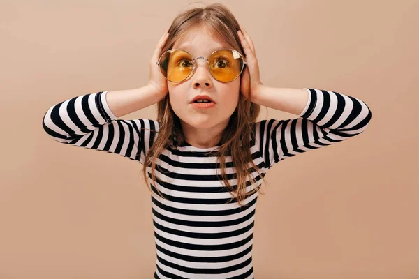 Adorável menina bonito vestindo redondo óculos amarelos parece preocupado, de mãos dadas na cabeça e olhando para longe — Fotografia de Stock