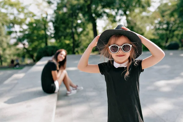 Κομψό μικρό κορίτσι φορώντας γυαλιά ηλίου και μαύρο φόρεμα που ποζάρει με την κάμερα με υπέροχο χαμόγελο με τη μητέρα στο παρασκήνιο — Φωτογραφία Αρχείου