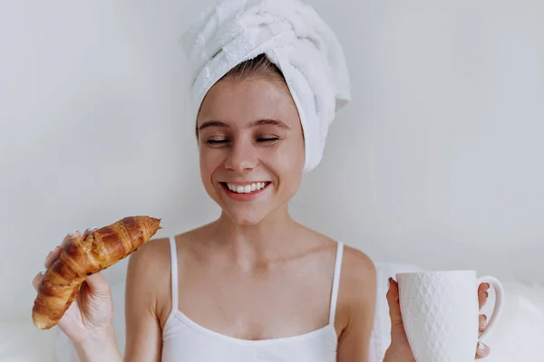 Muda bahagia tersenyum menarik wanita dengan handuk di kepala duduk di tempat tidur putih besar dan minum kopi dan makan croissant — Stok Foto