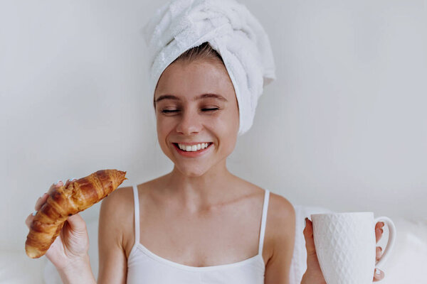 Young happy smiling attractive woman with towel on the head sitting in a big white bed and drinking coffee and eating croissant