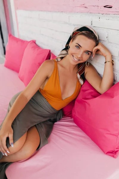 Portrait of laughing caucasian girl in orange swimsuit sits relaxedly with coffee on pink pillows on summer terrace — Stock Photo, Image