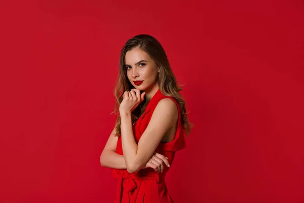 Wanita cantik yang percaya diri mengenakan gaun merah dengan bibir merah berpose ke kamera di atas latar belakang merah — Stok Foto