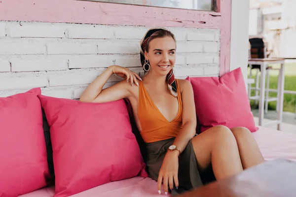 Retrato de senhora romântica elegante sentado no café com almofadas rosa bebendo café, vestindo camiseta laranja e saia — Fotografia de Stock