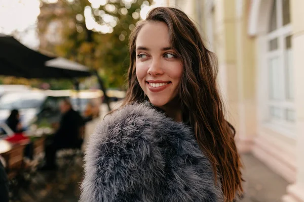 Gadis Kaukasia bergaya menarik dengan mantel bulu berpaling dan tersenyum di latar belakang kota. Sophisticated gadis menikmati musim gugur cerah pagi . — Stok Foto