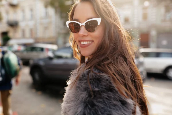 멋진 안경을 쓰고 모피 코트를 입고 도시 배경에서 햇빛을 받으며 카메라를 보고 있는 행복 한 미소짓는 여자 — 스톡 사진