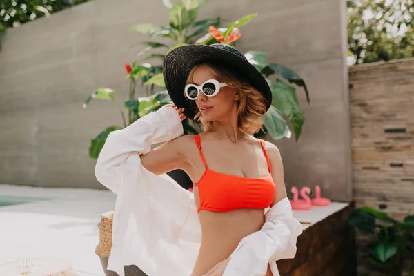 Mulher bonita elegante atraente vestindo chapéu preto, maiô laranja e camisa branca posando perto da piscina à luz do sol — Fotografia de Stock