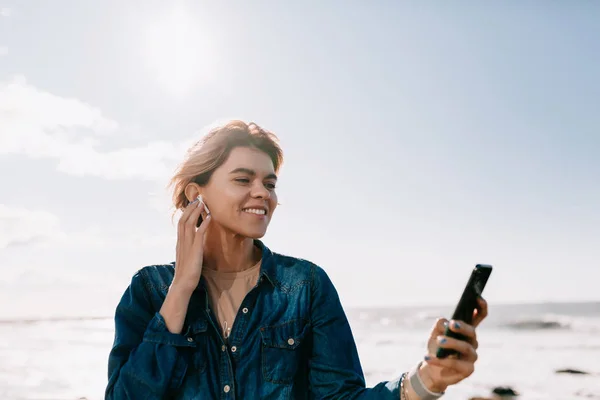 Захоплююча усміхнена дівчина розважається на пляжі вранці вихідних і позує на фоні неба зі смартфоном і слухає музику — стокове фото