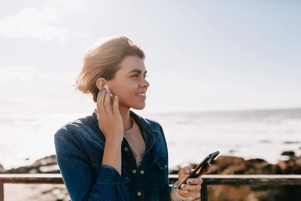 Щаслива жінка зі збиранням волосся в джинсовій сорочці слухає музику і насолоджується океаном і камінням в сонячний день — стокове фото