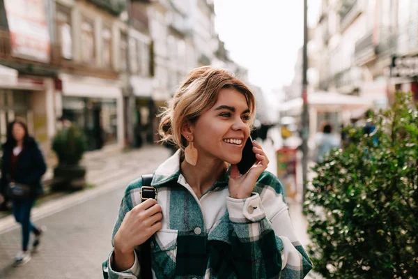 Wanita yang tersenyum gembira dengan rambut pirang yang dikumpulkan berbicara di telepon pintar dan menikmati perjalanannya di Eropa — Stok Foto