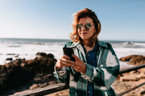 Зовнішній портрет привабливої жінки з кучерявою короткою зачіскою в смугастій сорочці з використанням смартфона на березі океану з камінням — стокове фото