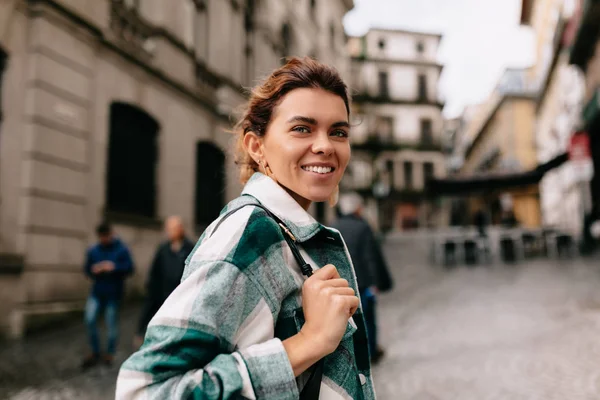 Happy tersenyum wanita dengan rambut pirang mengenakan kemeja bergaris berjalan di jalan tua yang cerah di Eropa . — Stok Foto