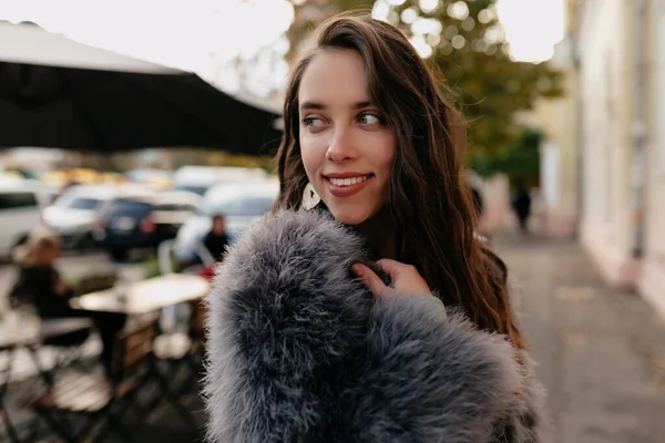 De cerca retrato exterior de la mujer encantadora tímida con el pelo oscuro y el uso de abrigo de piel mirando hacia otro lado y sonriendo — Foto de Stock