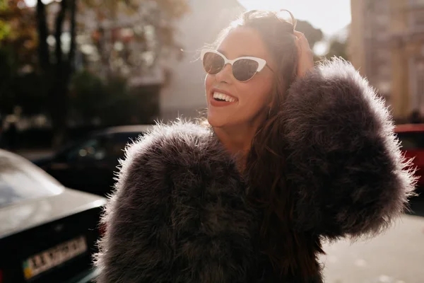 Sonriente joven atractiva con peinado oscuro con abrigo de piel y gafas elegantes sonriendo y divirtiéndose en la ciudad a la luz del sol —  Fotos de Stock