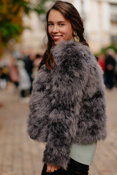 Привлекательная улыбающаяся европейская женщина с темными волнистыми волосами в стильной шубе, прогуливающаяся по городу в хороший день — стоковое фото
