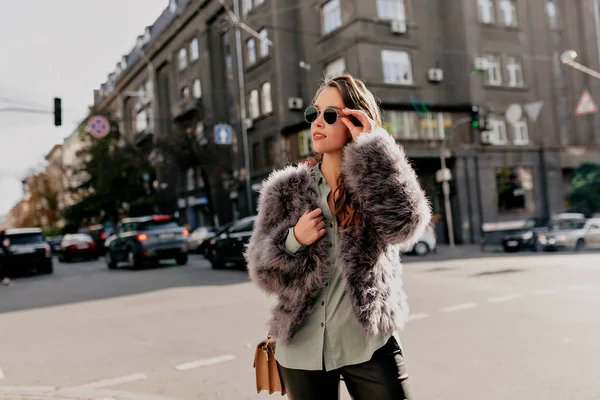 Adorable young lady with bag wearing fur coat and dark sunglasses posing on city background in sunny day. Happy woman spending time in city metropolis — 스톡 사진