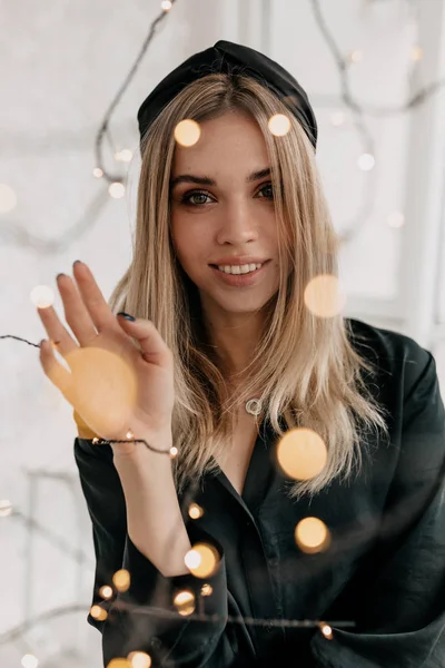 Retrato interior de la encantadora mujer rubia con una sonrisa encantadora con vestido negro y accesorios en la cabeza posando a la cámara con luces brillantes —  Fotos de Stock