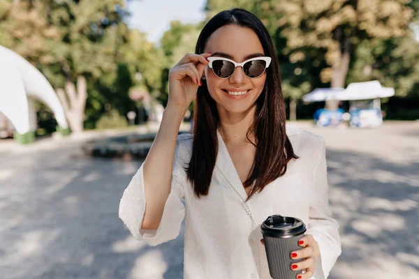 Happy smiling girl wearing white shirt and white glasses drinking coffee outside in good sunny day in the city park — 스톡 사진