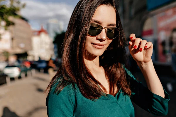 Di luar potret wanita european yang menawan mengenakan kacamata hitam dan blus hijau yang berpose di kamera di latar belakang kota di bawah sinar matahari — Stok Foto
