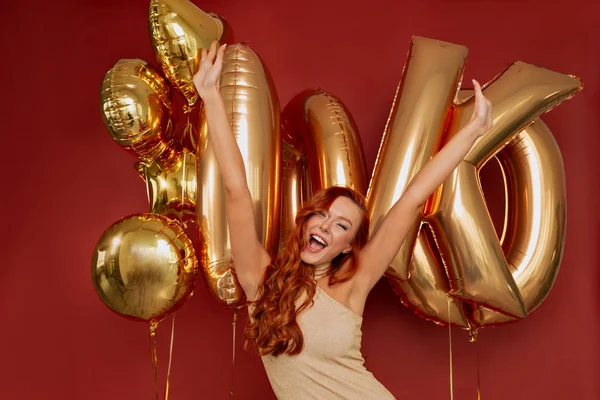 Festive girl in good mood wearing party dress having fun with raised up hands against background with balloons — Stock Photo, Image