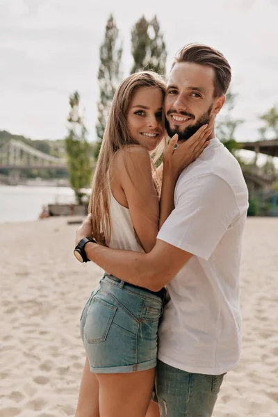 Pasangan muda yang manis dengan emosi bahagia berpelukan dan berpose di kamera selama pemotretan di pantai Stok Gambar Bebas Royalti