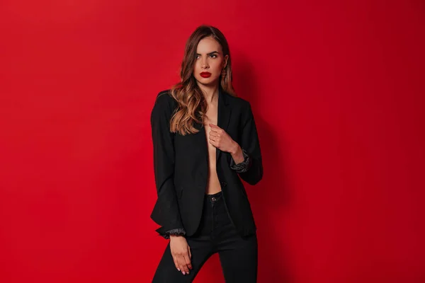 Elegante mujer confiada en traje negro posando sobre fondo rojo en el estudio —  Fotos de Stock