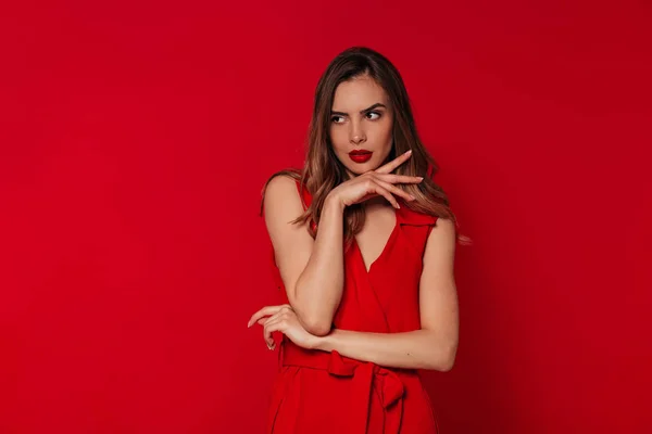 Välvillig kvinna med kväll smink bär röd klänning poserar över röd bakgrund i studio. Porträtt av europeisk självsäker kvinna — Stockfoto