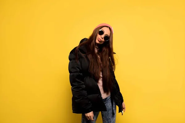 Стильная модная женщина в черной куртке, солнечных очках и розовой кепке позирует в студии на изолированном желтом фоне — стоковое фото