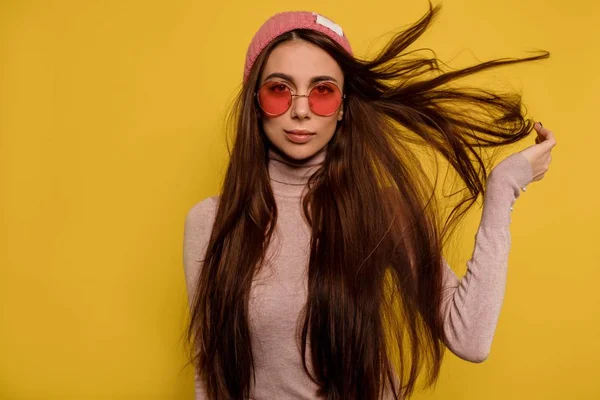Fashion portrait of showy girl with flying hair wearing round pink glasses and cap over isolated yellow background — 스톡 사진