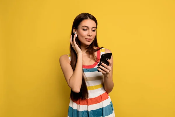 Wonderful woman with long hair and nude make up wearing bright dress listening music and holding smartphone over yellow background