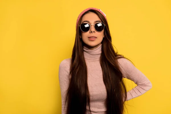 Indoor studio shot of trendy stylish woman with long dark hair wearing pink cap and black glasses over yellow background — 스톡 사진